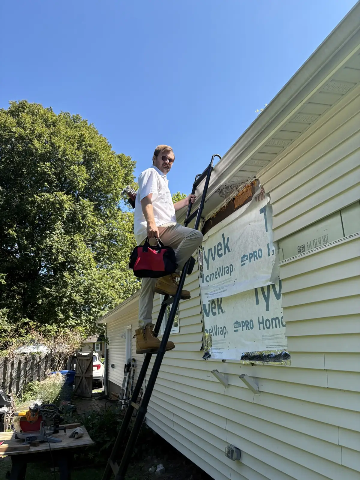 A technician with sunglasses and a red tool bag climbing a black ladder to the roofline