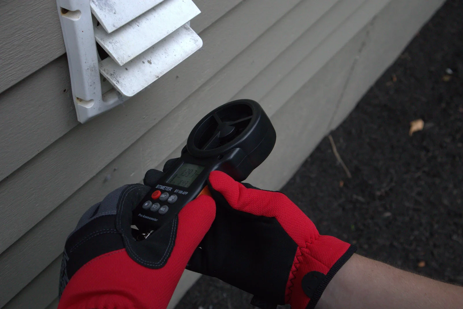 A gloved hand holding an airflow meter up to an exterior dryer vent hood