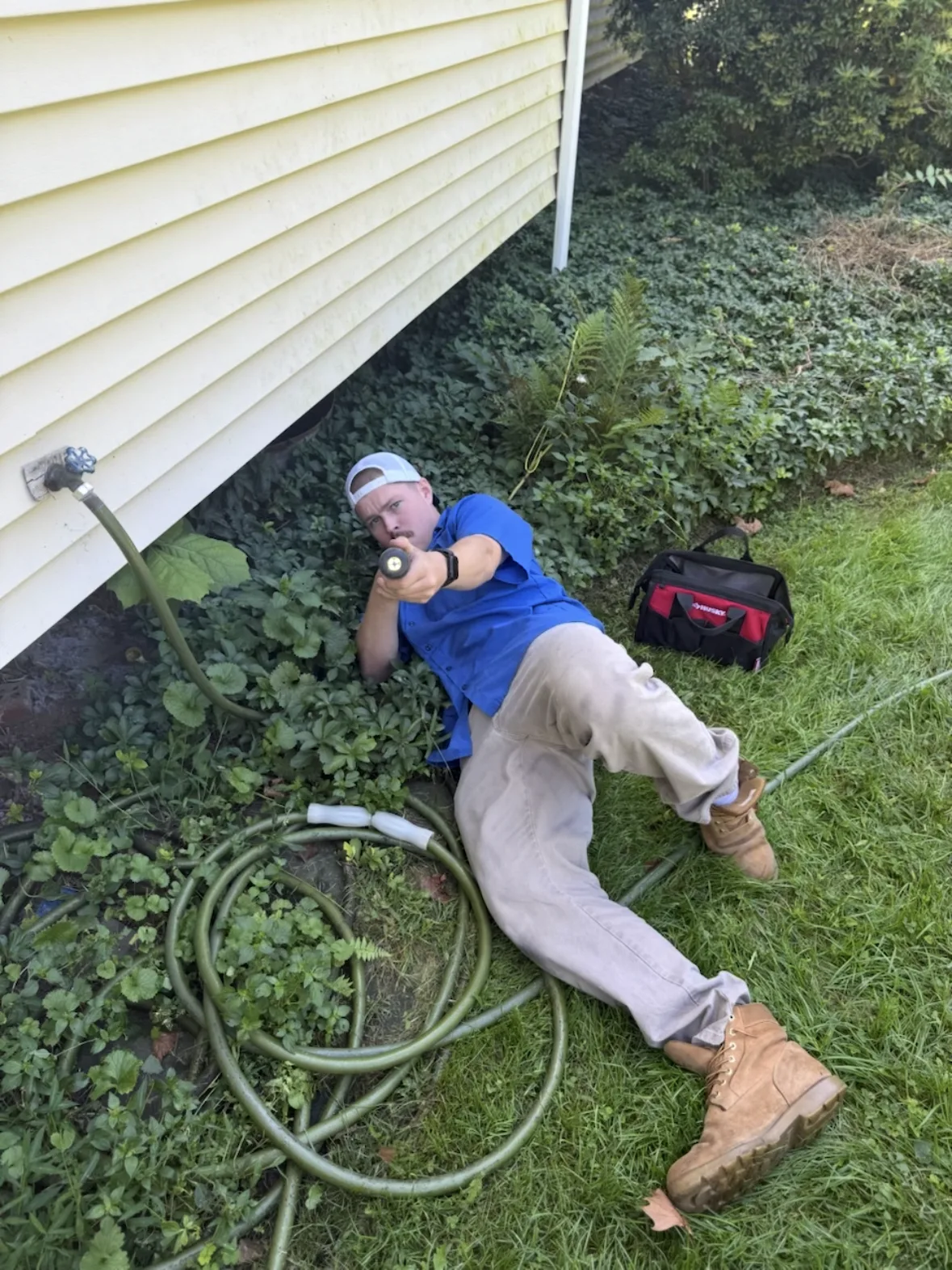 A Lint Guys technician on the ground outside a house pointing a flashlight at a dryer vent