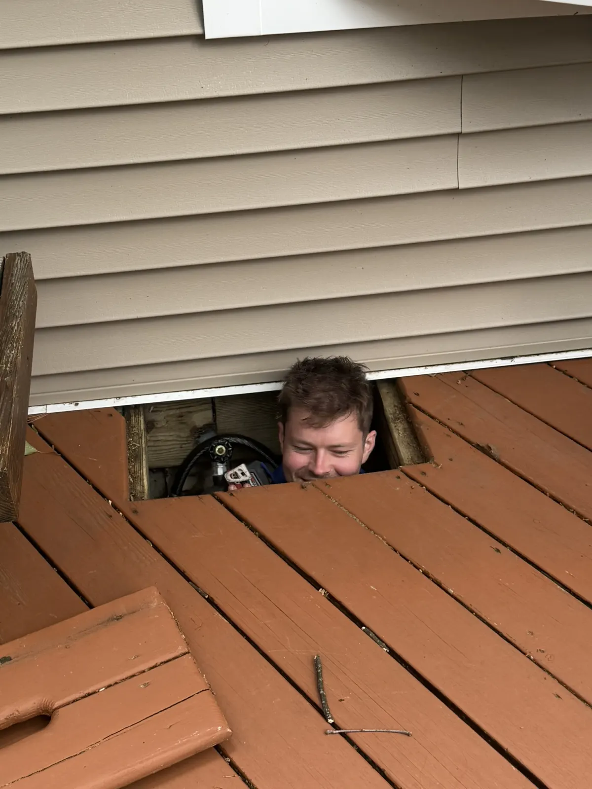 A Lint Guys technician looking up through an opening in a deck while reaching the dryer vent beneath it
