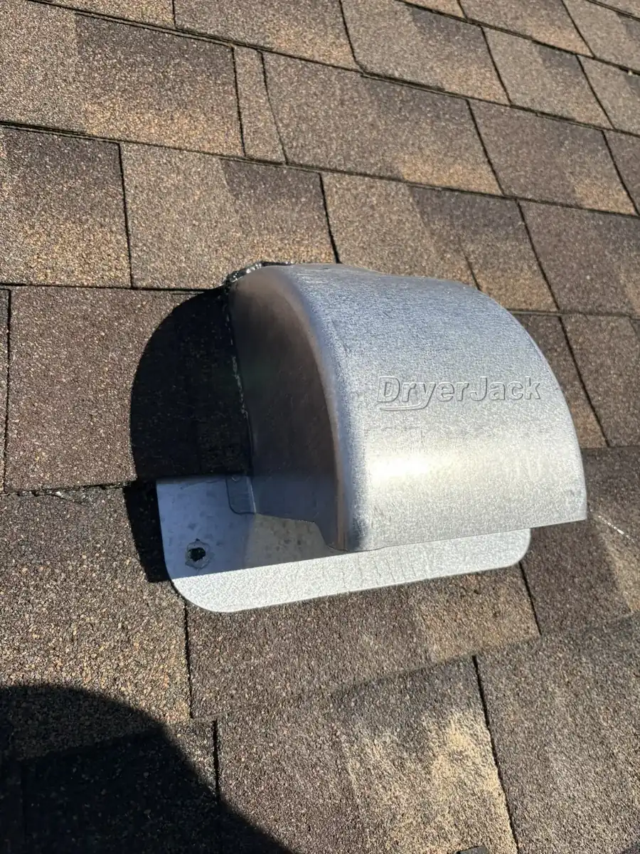 A galvanized hooded dryer vent on brown asphalt shingles in bright sun