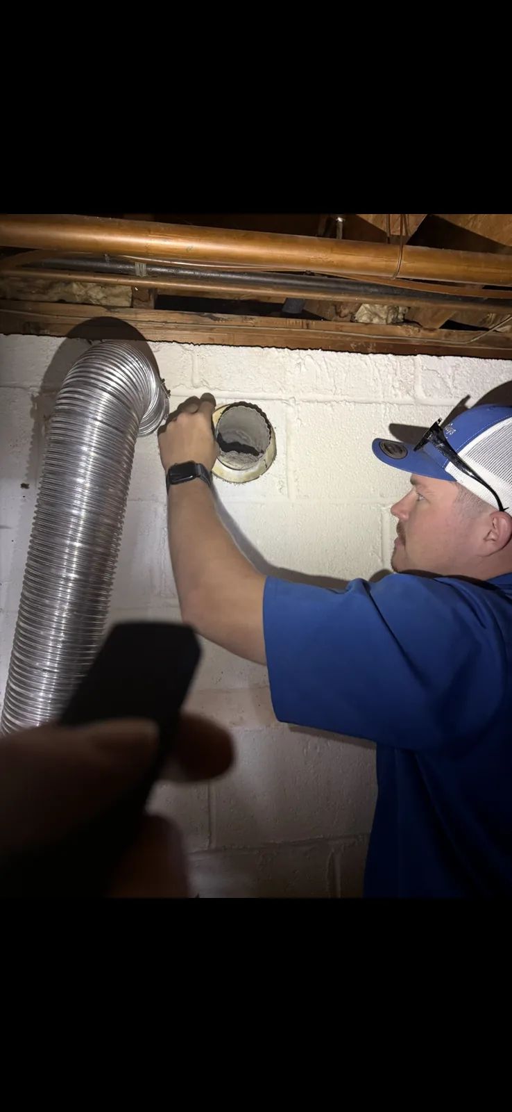A technician in a blue cap fitting flexible foil duct to a basement wall opening