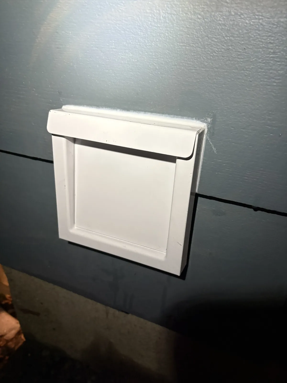 A square white flap-style dryer vent cover mounted on blue-grey siding